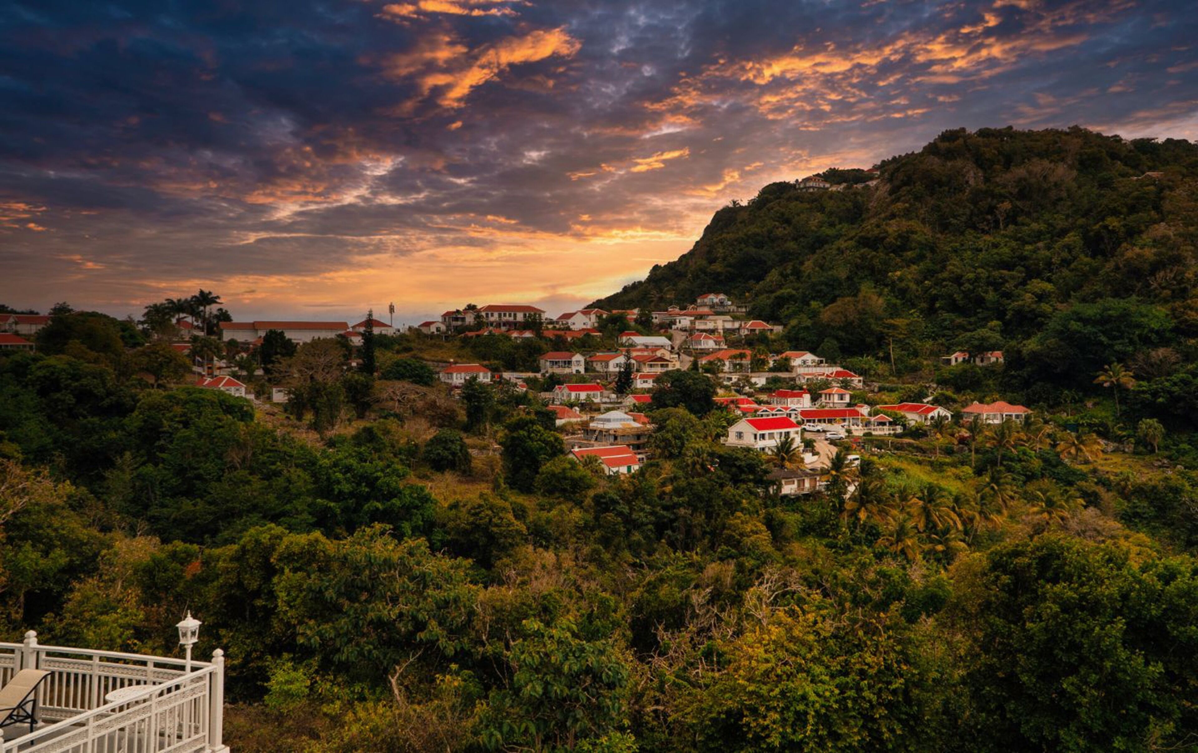 A hillside village with red-roofed houses, lush greenery, and a dramatic sunset sky.
