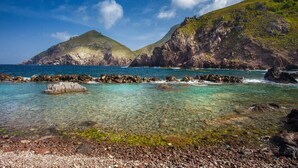 A coastal landscape with clear turquoise waters, rocky shoreline, and green hills.