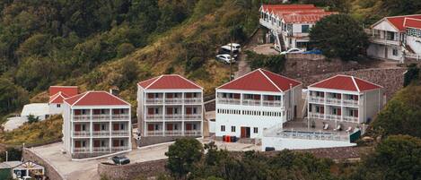A cluster of white buildings with red roofs, each featuring multiple balconies, situated on a hillside with dense greenery around.