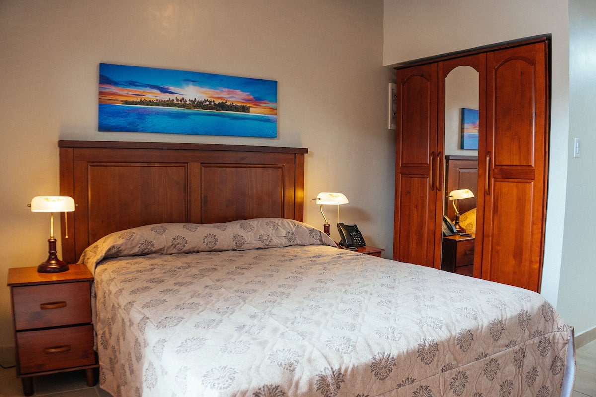 Una cama bien hecha con una colcha estampada con motivos florales, dos lámparas de noche, una mesita de noche de madera, un armario de madera y un cuadro de un atardecer sobre el agua en la pared.