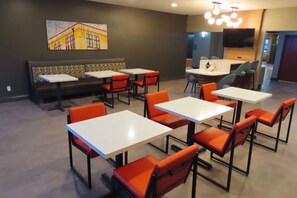 A modern dining area with white tables and orange chairs, a large painting on the wall, and a TV mounted on the wall.