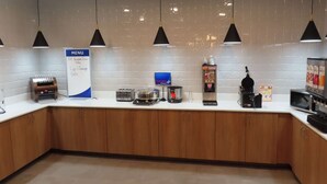 A modern kitchen with a white countertop, wooden cabinets, and black pendant lights. A menu board is visible, and various appliances like a coffee maker and microwave are present.
