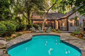 A swimming pool with stone tiles, surrounded by greenery and a house with string lights. - Your Private Clubhouse with Golf Simulator and Pool (Pinehurst)