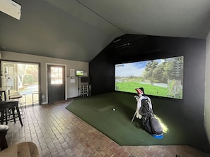 A room with a large screen displaying a golf course, a golf bag, and a bar area with stools. - Your Private Clubhouse with Golf Simulator and Pool (Pinehurst)