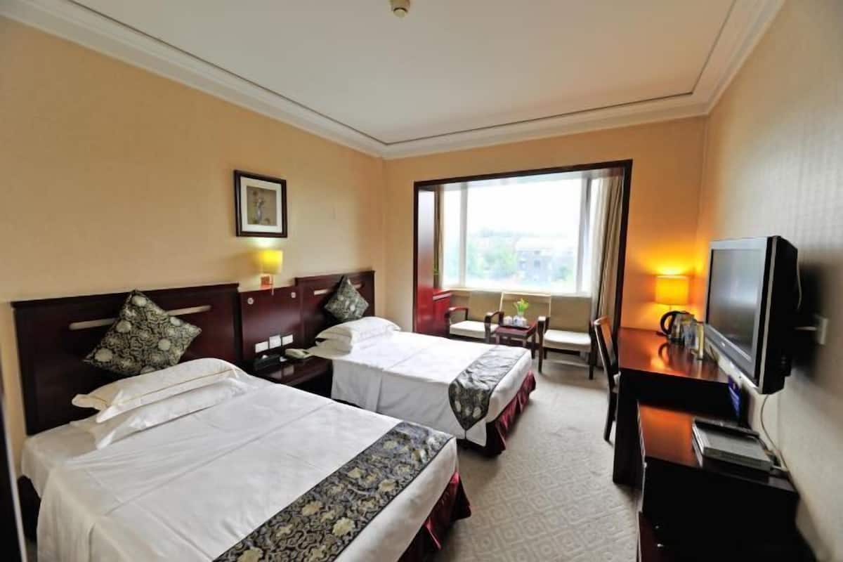 A hotel room with two beds, a desk with a TV, a window with a view, and a small table with a lamp.