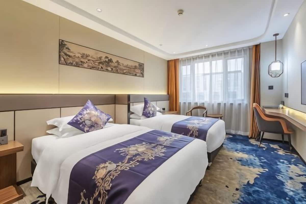 A hotel room with two beds, a desk, a chair, a large window with curtains, and a painting on the wall.