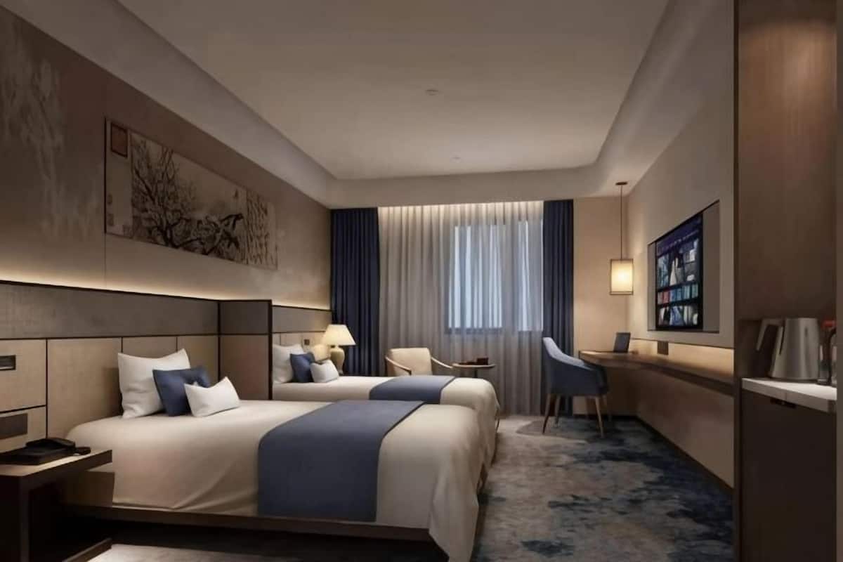 A hotel room with two beds, a desk, a chair, a lamp, and a large window with curtains.