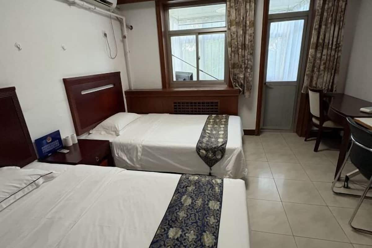 A hotel room with two beds, a desk, a chair, a window with curtains, and a heater.