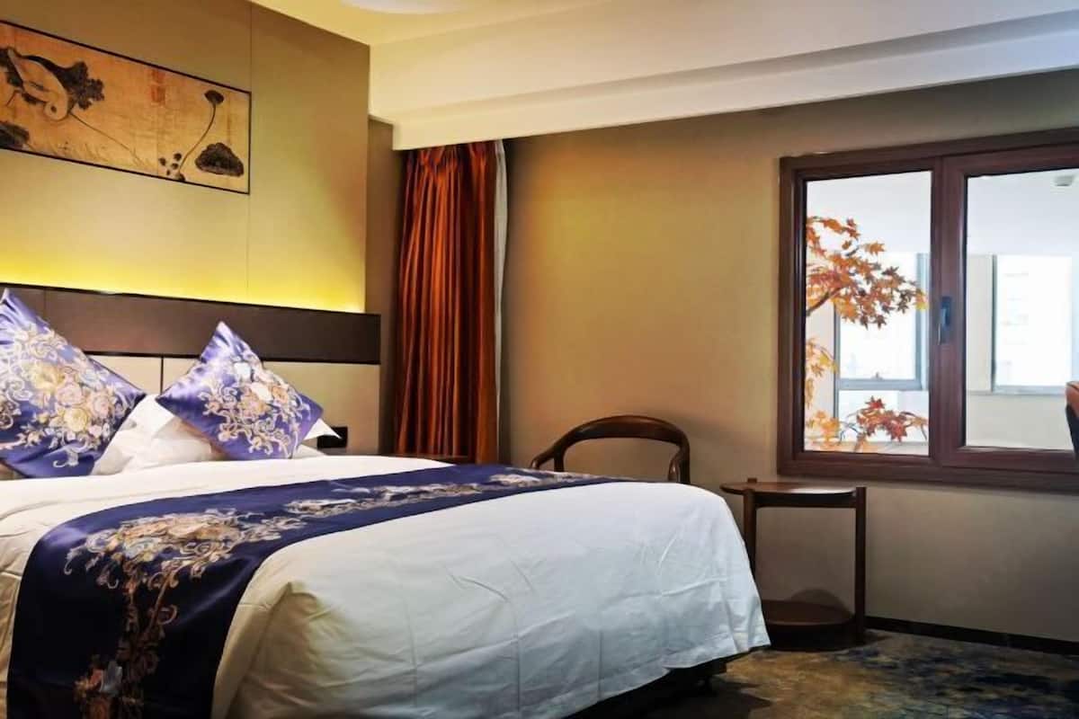 A hotel room with a bed, a window with autumn leaves, a chair, and a painting on the wall.
