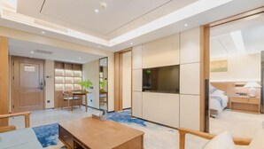 A modern hotel room with a wooden dining table, a sofa, and a TV. There is a bookshelf and a plant in the corner. - Beijing Post & Telecom Conference Centre (Beijing)