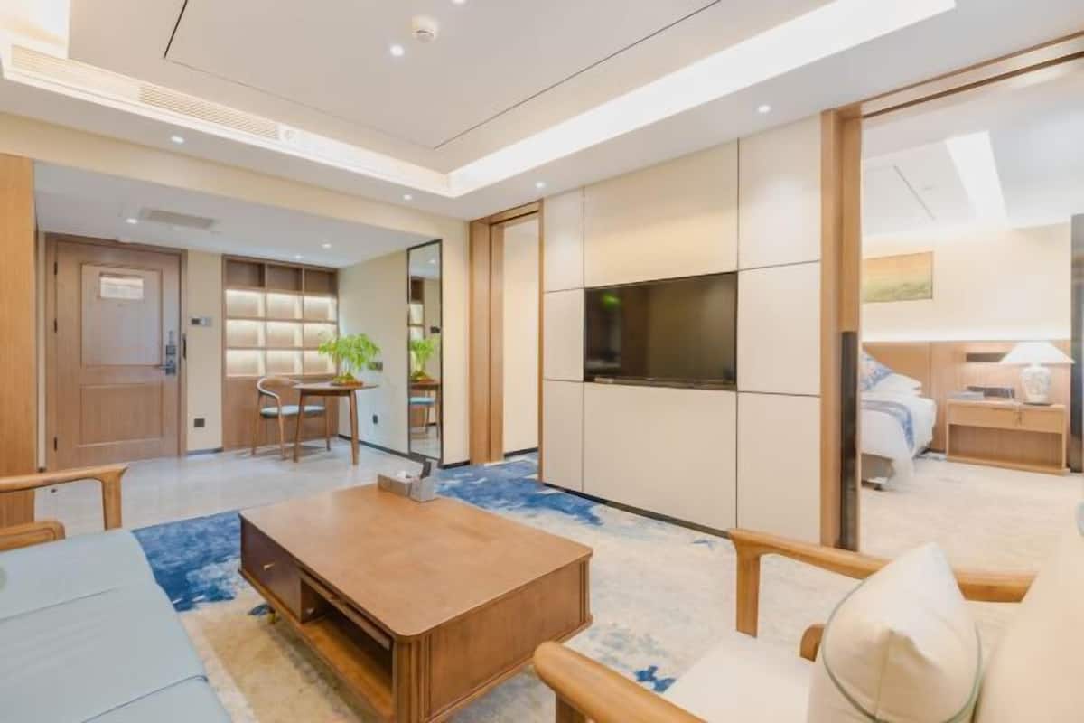 A modern hotel room with a wooden dining table, a sofa, and a TV. There is a bookshelf and a plant in the corner.