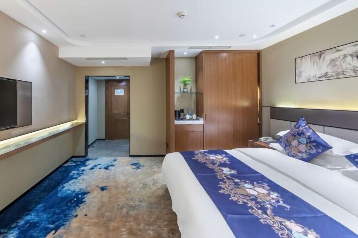 A hotel room with a bed, a TV, a wardrobe, and a desk with a plant.