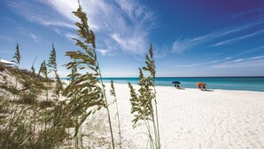 En strand med klar himmel, vit sand och högt gräs. Man kan se två parasoller och en strandstol.