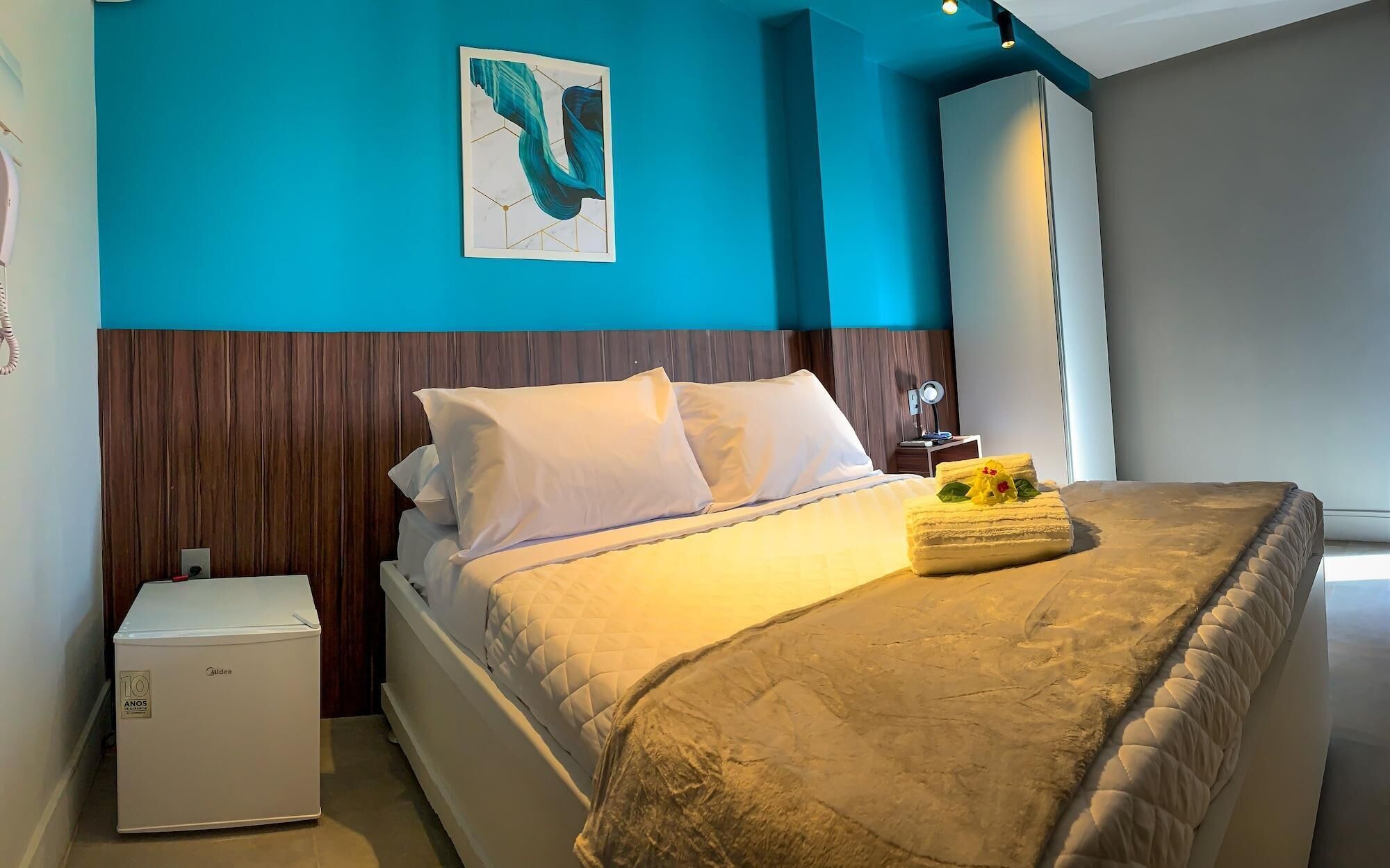 A hotel room with a bed, a nightstand, a small white appliance, and a painting on the wall.