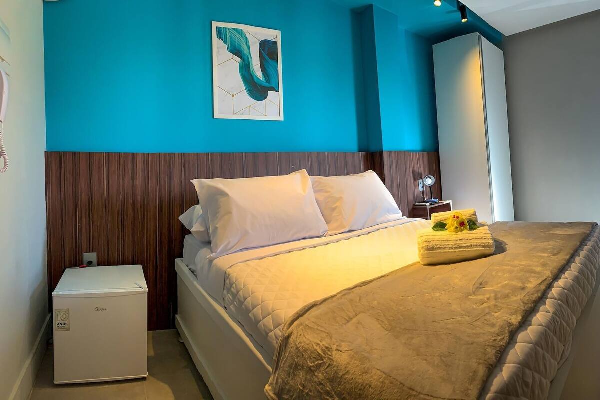 Una habitació d'hotel amb un llit, una tauleta de nit, un aparell petit i blanc, i un quadre a la paret.