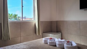 A hotel room with a large bed, a flat-screen TV mounted on the wall, a window with curtains, and a view of palm trees and buildings outside. - Hotel Atlântico Sul (Ipojuca)