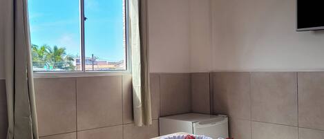 A hotel room with a large bed, a flat-screen TV mounted on the wall, a window with curtains, and a view of palm trees and buildings outside.