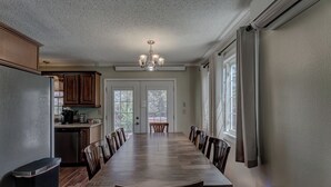 Ein Esszimmer mit einem langen Holztisch, umgeben von Stühlen, einer Kristallleuchtre und einer Küchenzeile mit Schränken und Geräten.