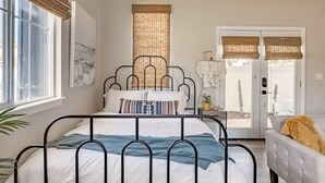 A bedroom with a metal bed frame, white bedding, a large window with blinds, and a view of the outdoors.