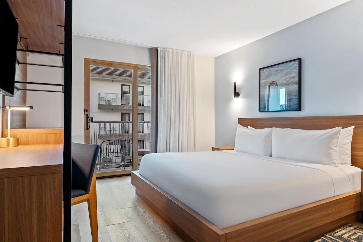 Habitación de hotel con cama de madera, un escritorio, una silla, un ventanal grande y un balcón con vistas a la ciudad.