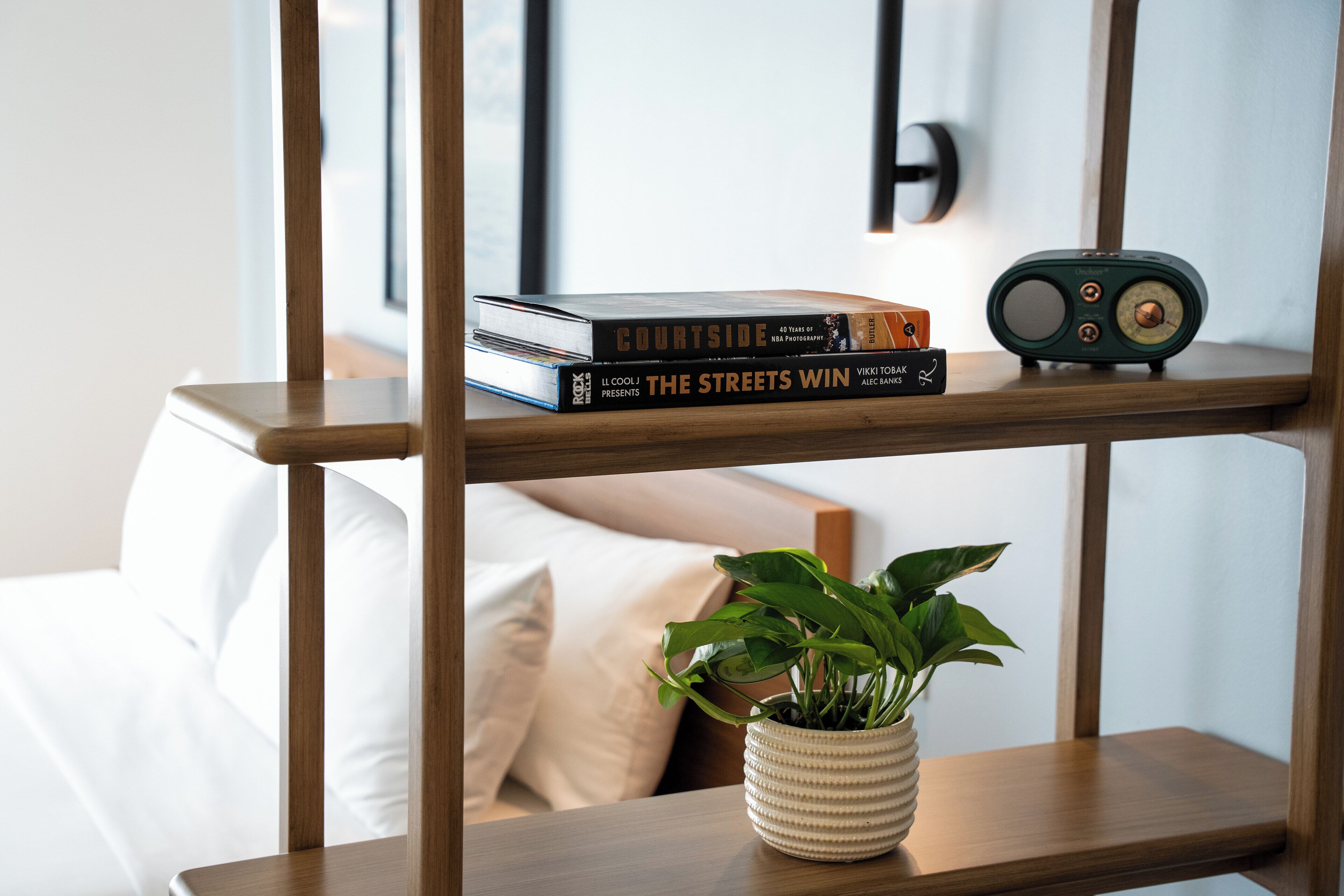A wooden shelf with books and a radio, a potted plant, and a bed with white bedding.