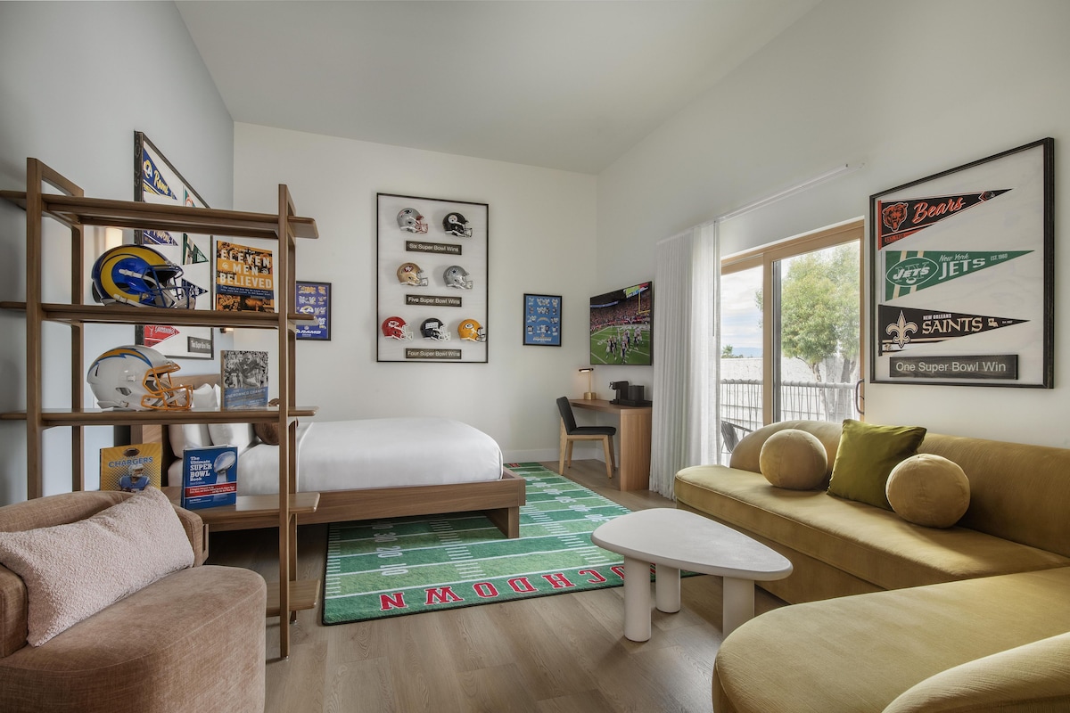 Una habitación de hotel moderna con una cama, un sofá, un escritorio y una estantería con recuerdos deportivos.