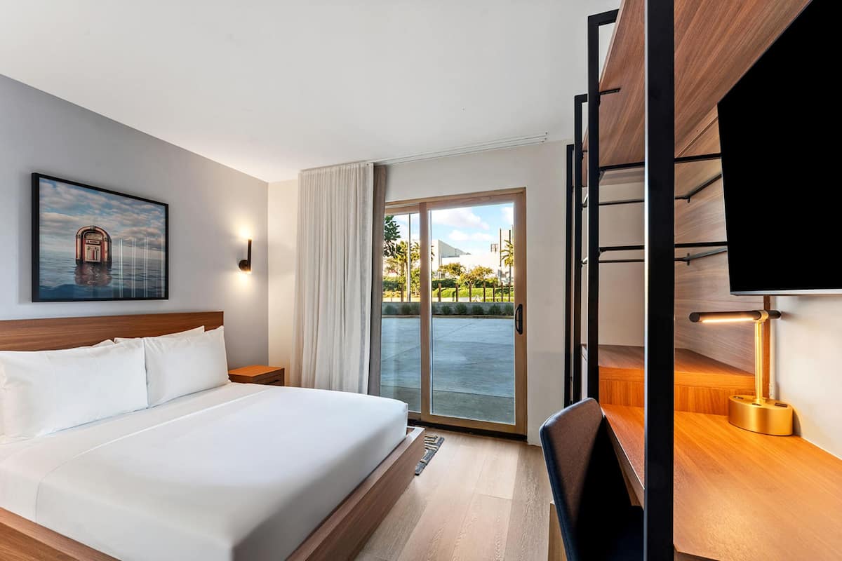 Habitación de hotel con cama, escritorio con lámpara, televisor, ventana con cortinas y un cuadro en la pared.