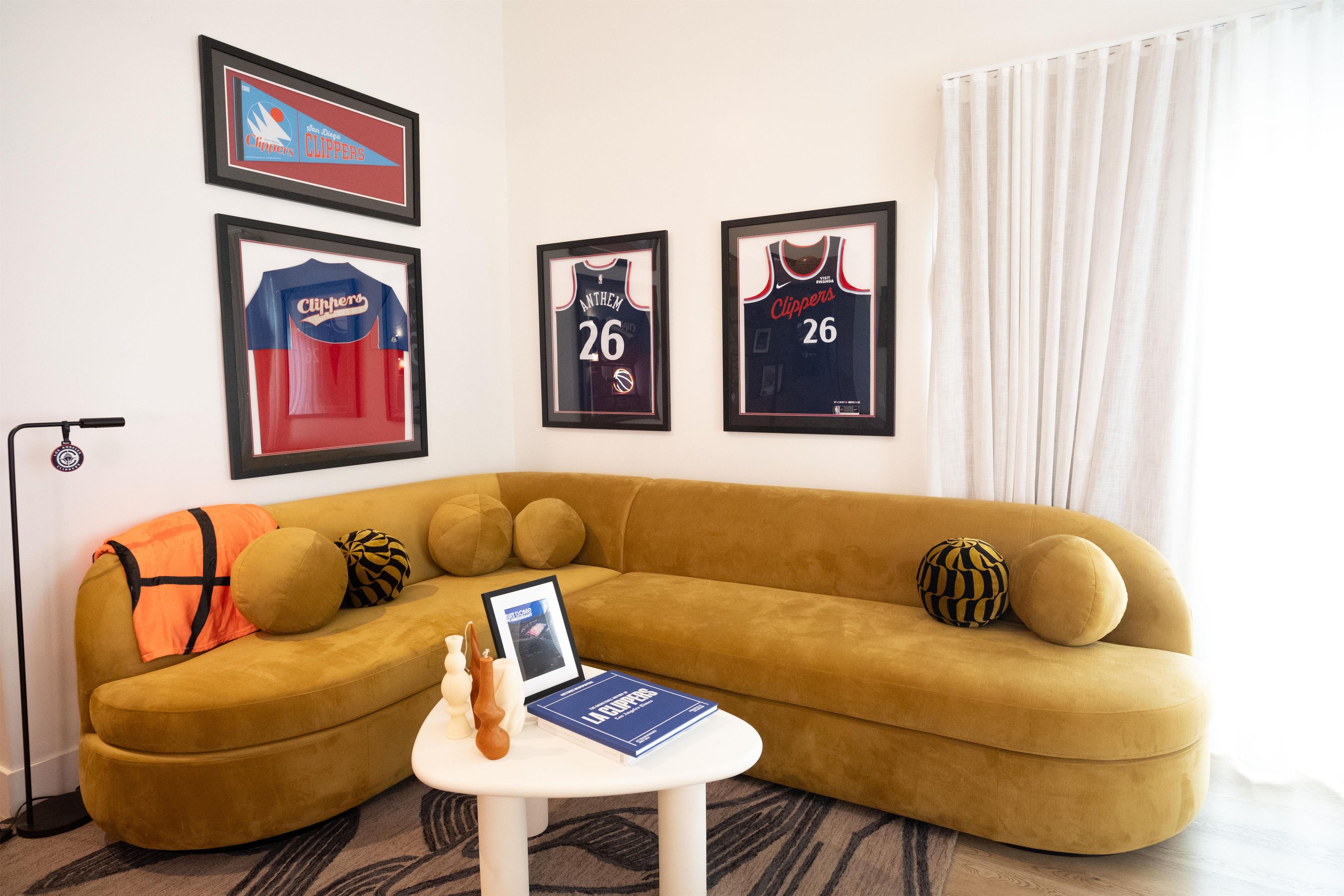 A living room with a mustard-colored sofa, a small white table with a book, and three framed sports jerseys on the wall.