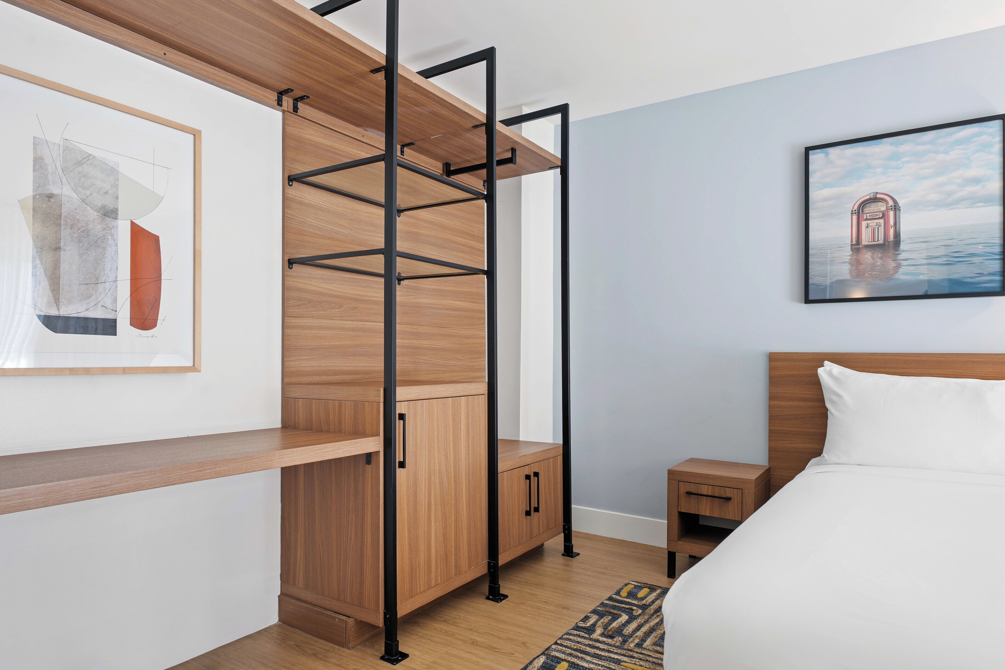 Un dormitorio con una cama, una mesita de noche de madera, una pequeña mesa, un estante en la pared y un cuadro.