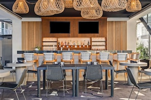 A modern bar area with wicker pendant lighting, wooden panel walls, and a variety of bottles and glasses on shelves. - The Anthem Los Angeles Stadium District, Tapestry by Hilton (Inglewood)