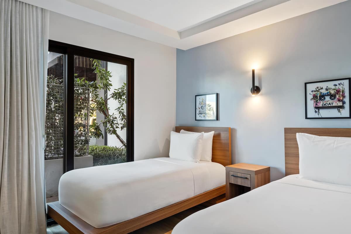 Habitación de hotel con dos camas, una ventana que da a zonas verdes, una lámpara de pared e imagen enmarco.