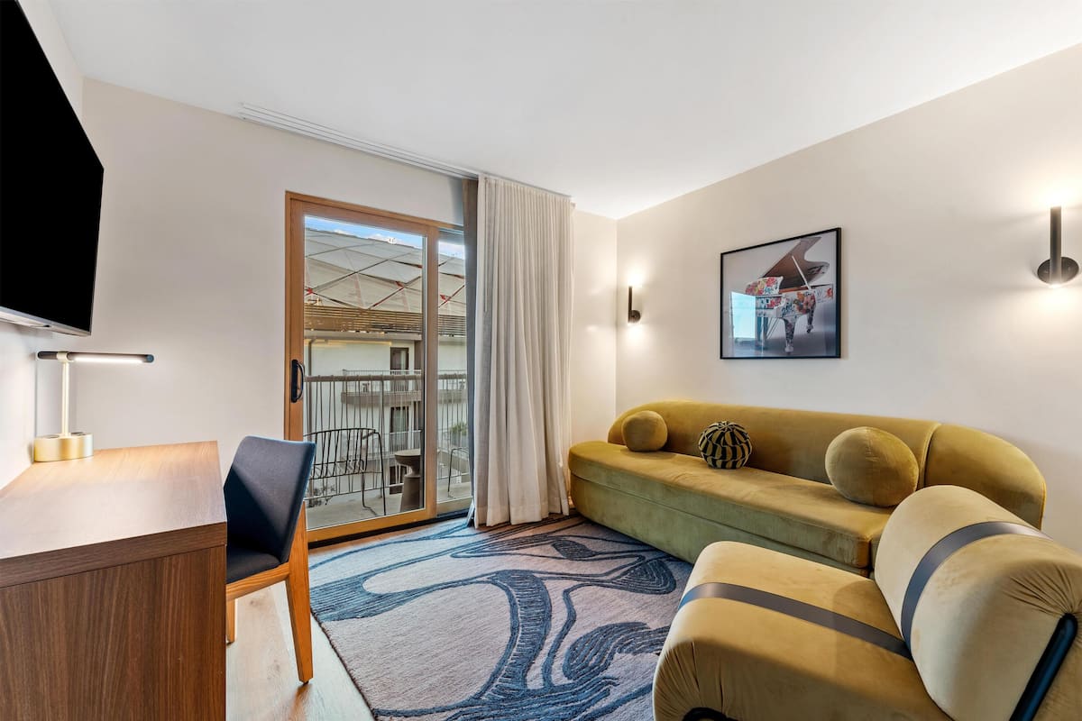 Una sala de estar moderna con un sofá, un escritorio y un televisor. Se aprecia una ventana grande con cortinas, una puerta que da a un balcón y un cuadro colgado en la pared.