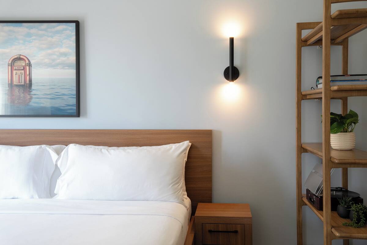 Una cama bien hecha con sábanas blancas, una mesita de noche de madera con un cajón, una lámpara de pared y un cuadro enmarcado de un edificio sobre el agua.