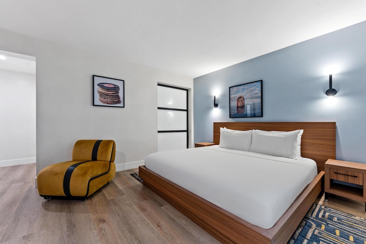 Una habitación de hotel moderna con una cama grande, una mesita de noche de madera, un sillón amarillo y dos lámparas fijadas a la pared.