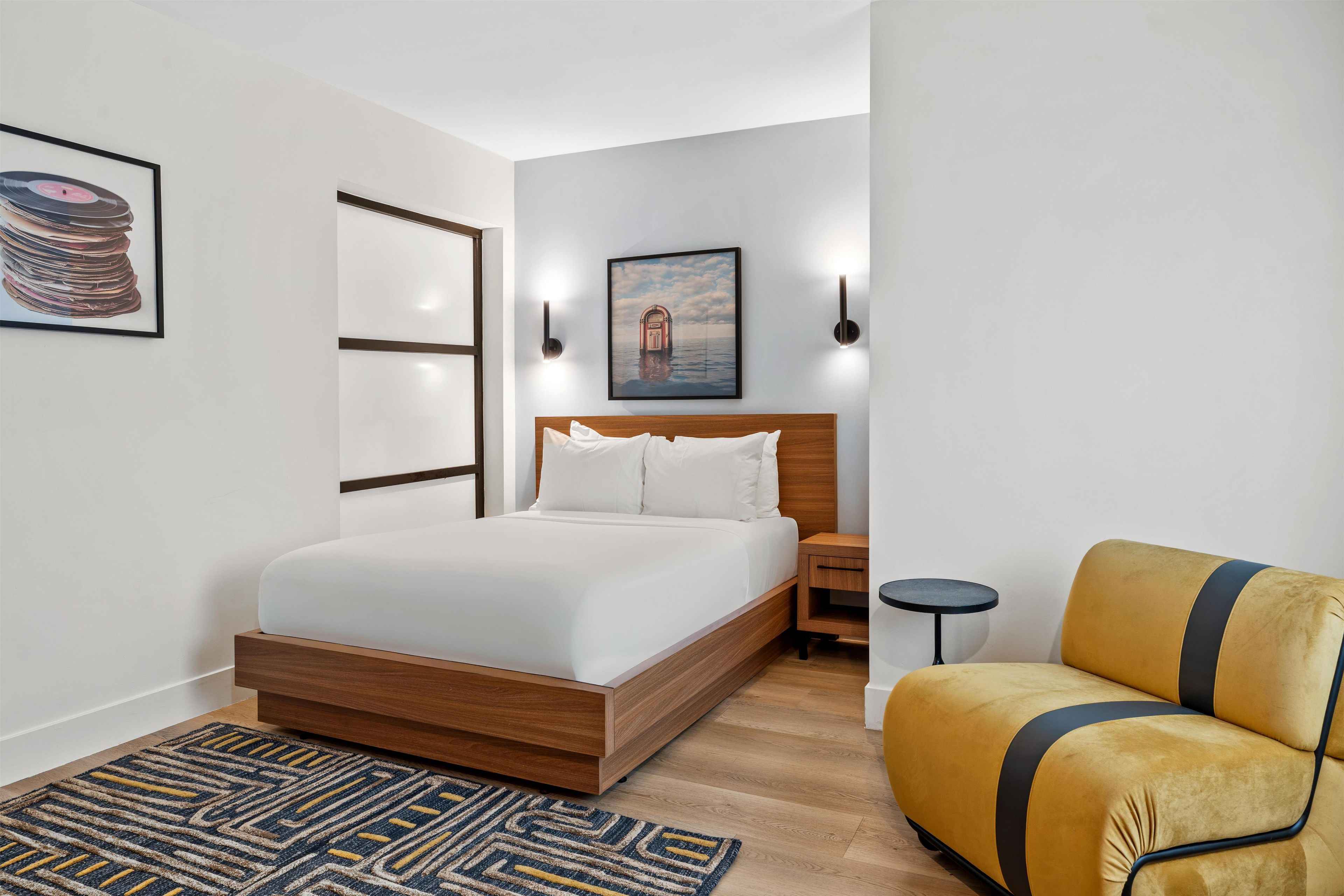 A modern bedroom with a wooden bed, a small bedside table, a round nightstand, a yellow sofa, a patterned rug, and two wall-mounted lights.