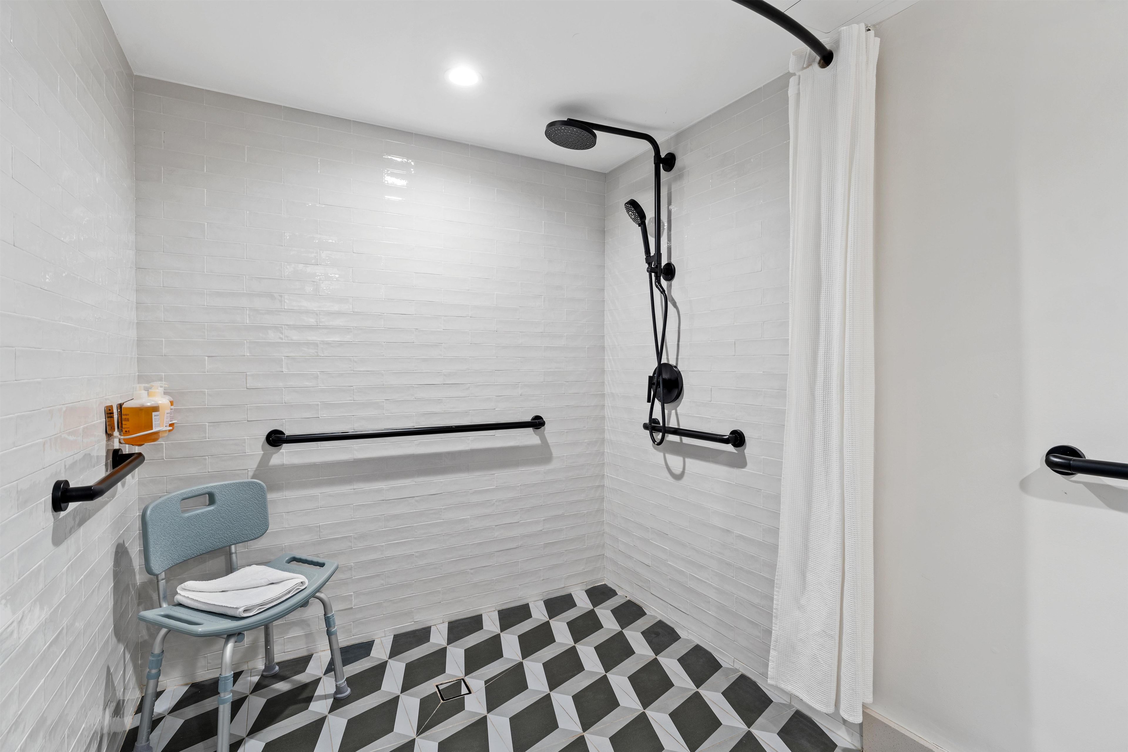A bathroom with white tiled walls, a black and white checkered floor, a wheelchair-accessible chair, and a shower with a handheld showerhead.