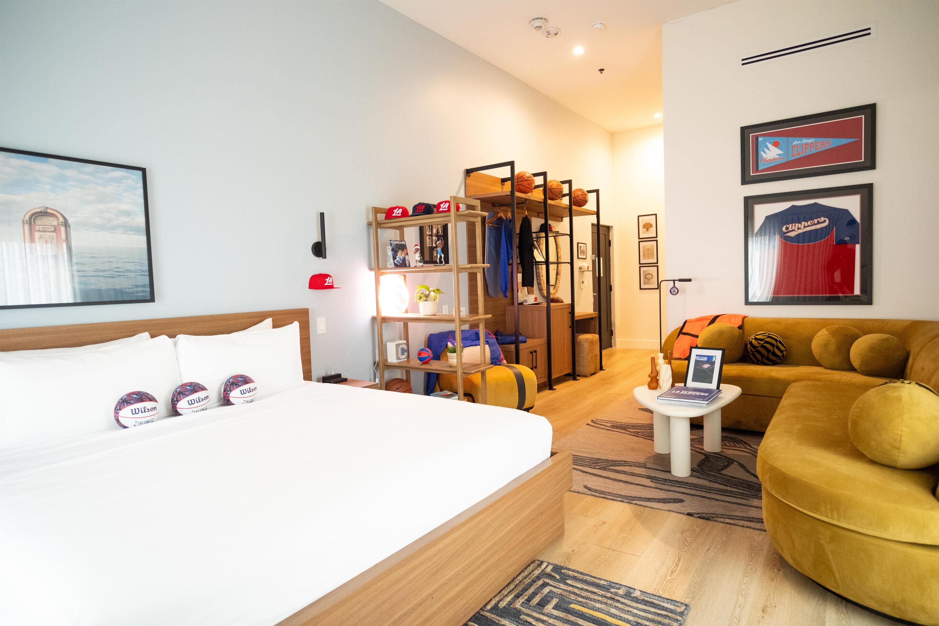 A hotel room with a bed, a sofa, a small table, a bookshelf, and framed sports jerseys.