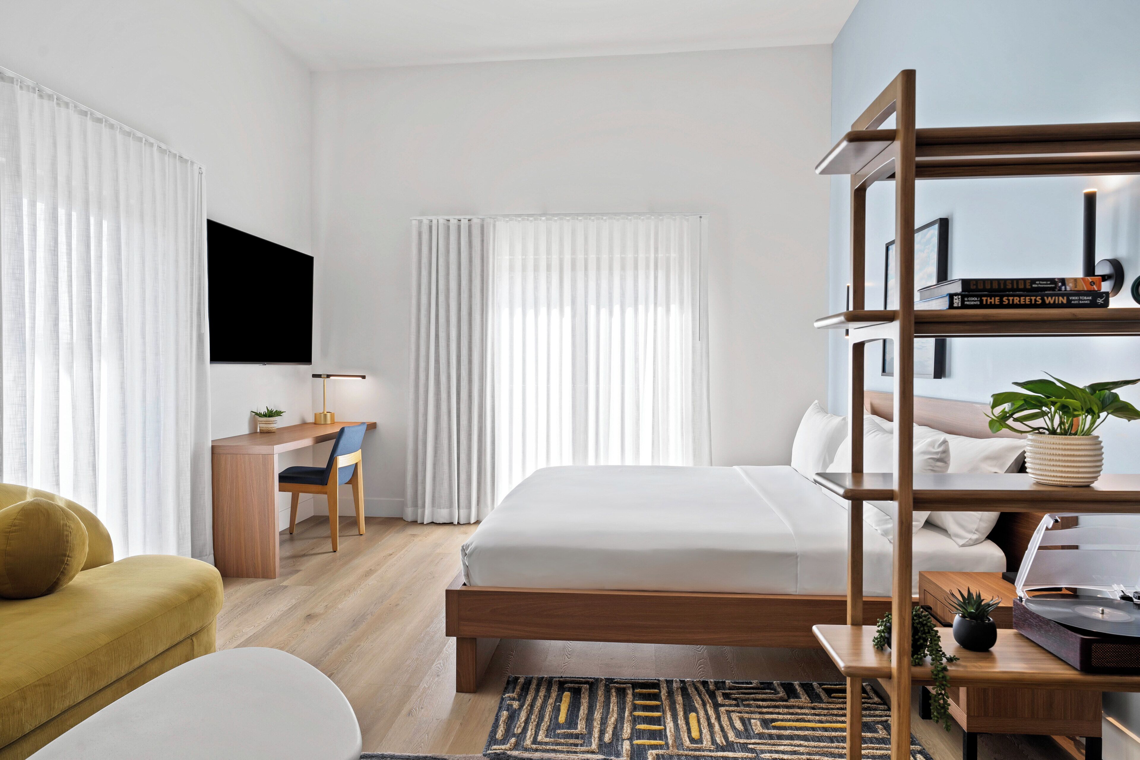 A modern hotel room with a wooden bed, a desk, a chair, a television, and a bookshelf.