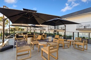 Un área de estar al aire libre con mobiliario de madera y grandes sombrillas, con vistas a un edificio moderno con ventanales amplios.