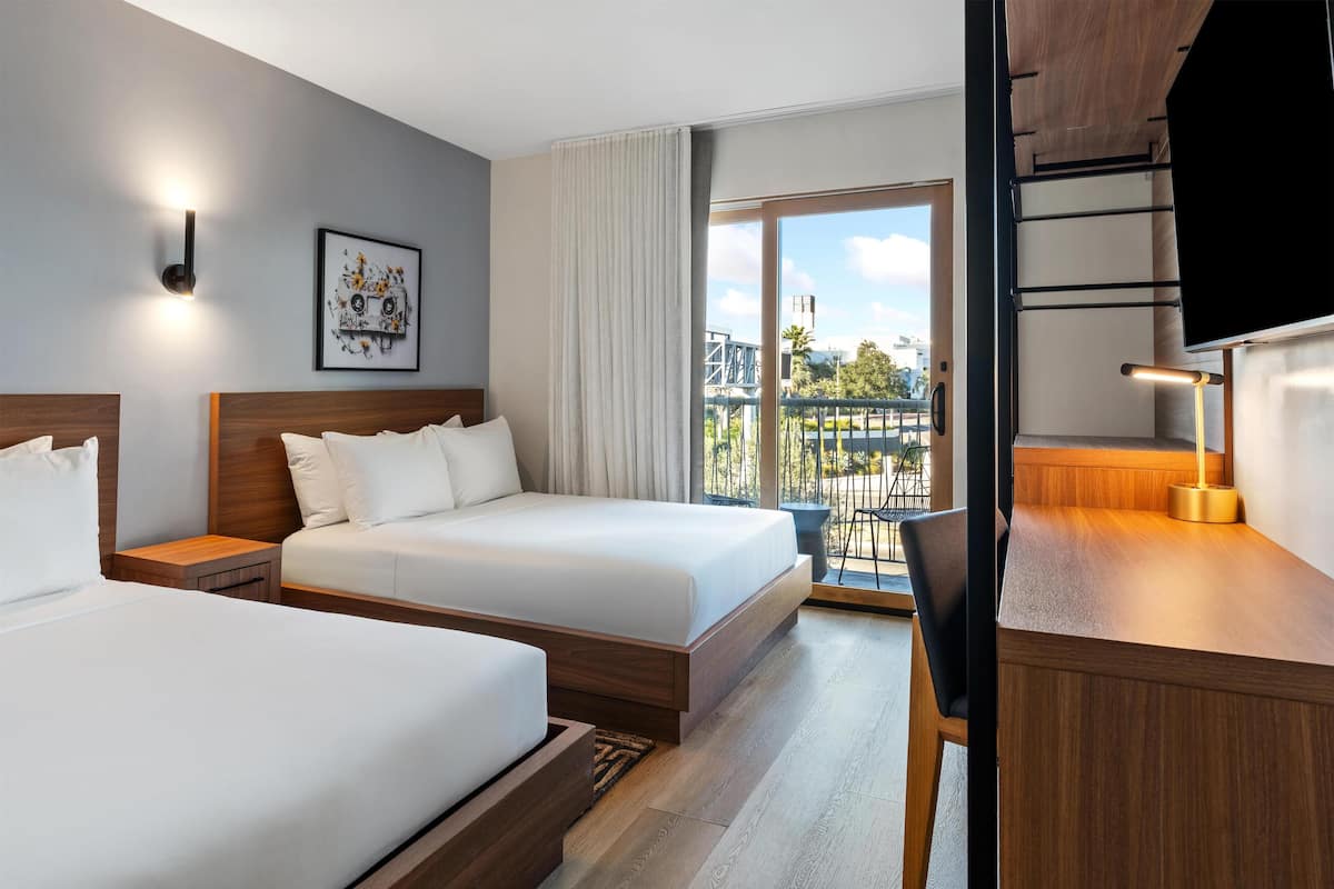 Habitación de hotel con dos camas, un escritorio, una silla, un televisor y un balcón con vistas a la ciudad.