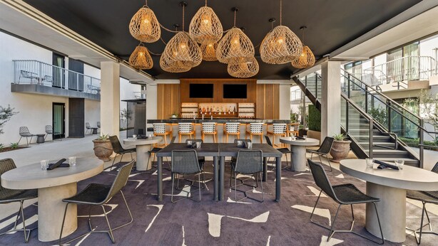 Un área de comedor exterior moderna con mesas y sillas redondas, una barra con taburetes y una escalera que conduce a un nivel superior.