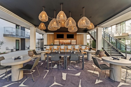 A modern outdoor dining area with round tables and chairs, a bar with stools, and a staircase leading to an upper level.
