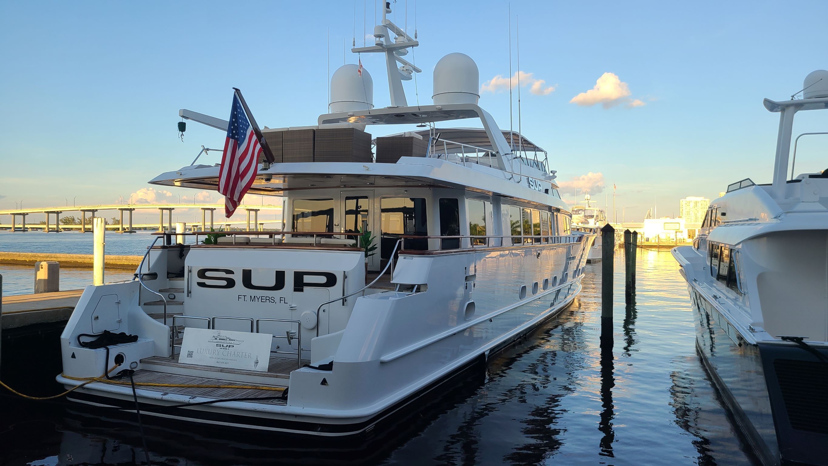 A luxury yacht docked at a marina with a clear view of the water and a bridge in the background.