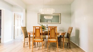 Un comedor con una mesa de madera y cuatro sillas de mimbre, una lámpara de araña y un espejo enmarcado en la pared.