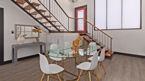 Un espace repas moderne avec une table en verre et des chaises blanches, un escalier avec des marches en bois et des rampes noires, et une fenêtre à cadre rouge.