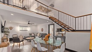 Un vaste espace de vie comprenant une table à manger en verre, des chaises blanches, un canapé gris et un escalier en bois.