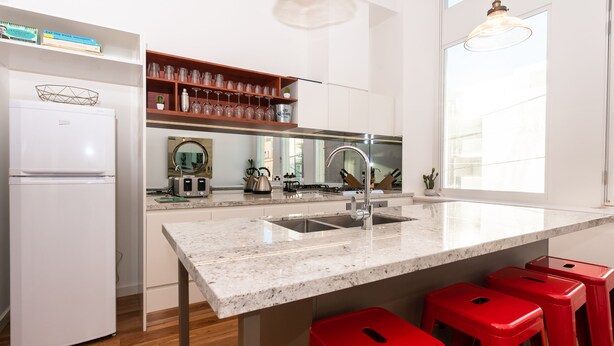 A modern kitchen with a white refrigerator, a built-in oven, a stainless steel sink, and a granite countertop. There are red bar stools and a window providing natural light. - Slick Home, Free Parking (Brunswick)