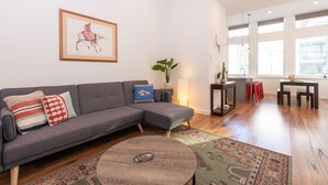 A modern living room with a grey sectional sofa, a round coffee table, and a colorful rug. There is a framed painting on the wall and a potted plant on a side table. - Slick Home, Free Parking (Brunswick)