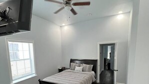 A bedroom with a bed, a ceiling fan, a TV mounted on the wall, and a window.