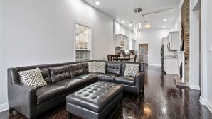 A modern living room with a sectional sofa and ottoman, a dining area, and a kitchen with white cabinets.